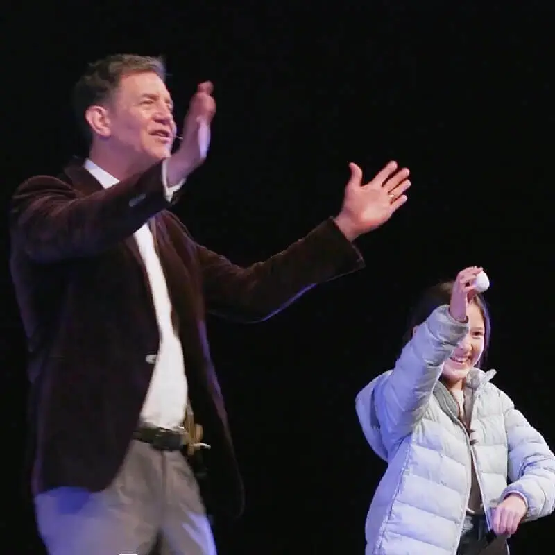 Doug performing with an audience volunteer. The volunteer smiles and holds an egg.